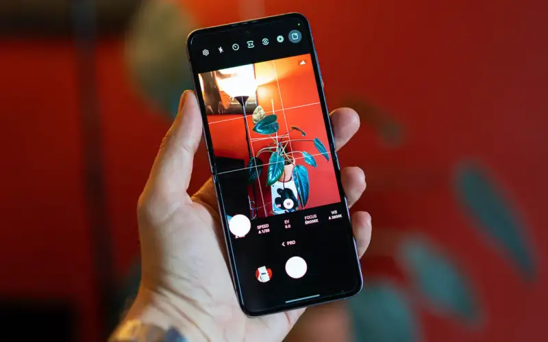 Dłoń trzyma smartfon z włączonym trybem PRO aparatu. Na ekranie widoczny jest kadr z rośliną w doniczce i lampą.