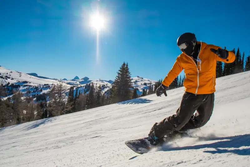 Snowboardzista w pomarańczowej kurtce zjeżdża po ośnieżonym stoku w słoneczny dzień.
