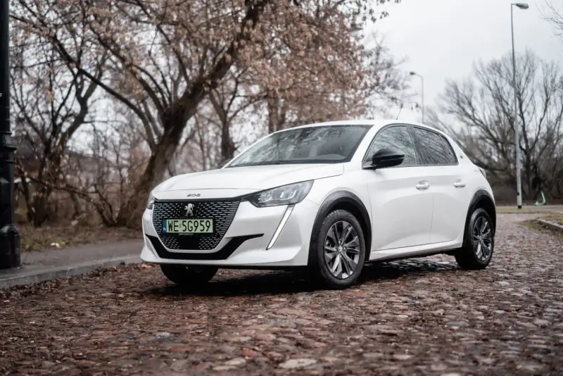 Biały Peugeot e-208 na brukowanej ulicy, z drzewami w tle.
