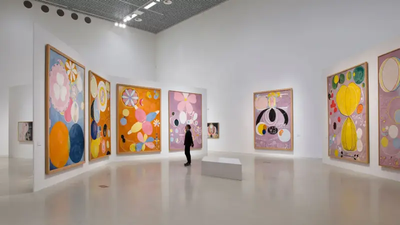 En person betraktar en samling färgstarka, abstrakta målningar av Hilma af Klint i ett ljust galleri.