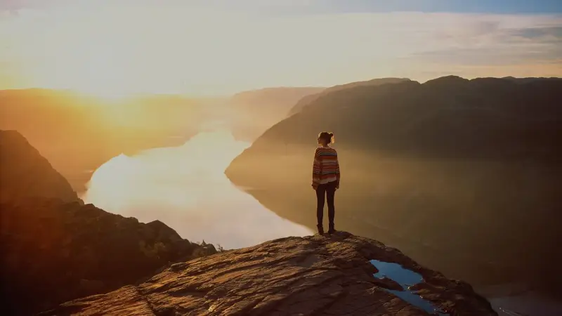 A lone figure stands on a cliff edge, gazing at a serene fjord bathed in golden sunset light. It's a moment to give it to God.