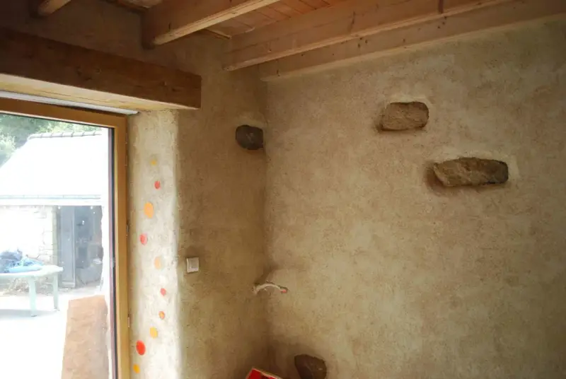 Un mur avec un enduit chaux finition texturé, intégrant des pierres apparentes et des touches de couleur. Le bois des poutres et du cadre de fenêtre apporte de la chaleur.
