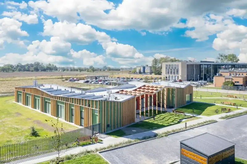 Nowoczesny budynek użyteczności publicznej z zielonymi fasadami i drewnianymi elementami, otoczony trawnikami i parkingiem.