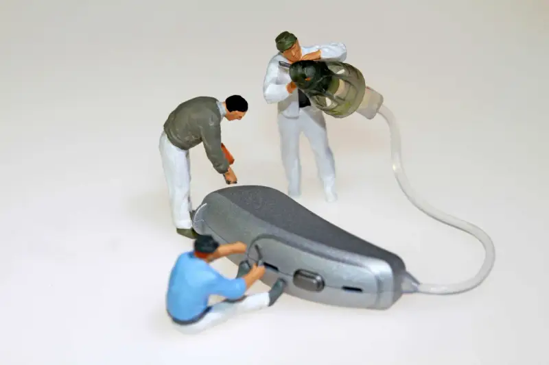 Miniaturowi pracownicy zajmują się naprawą aparatów słuchowych. Jeden z nich przykręca element, drugi czyści słuchawkę, a trzeci pracuje nad aparatem.