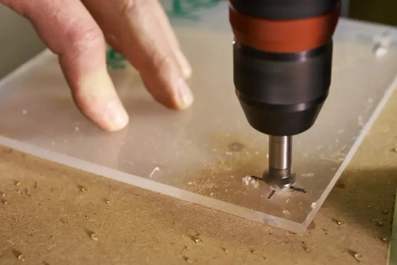 A drill bit is used to drill through acrylic. A hand guides the acrylic sheet as the drill creates a hole, showing it's possible to drill through acrylic.