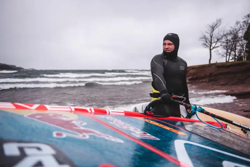 Mann in Neoprenanzug mit Windsurfbrett am Strand. Kaltes Wetter für Windsurfen in Deutschland.