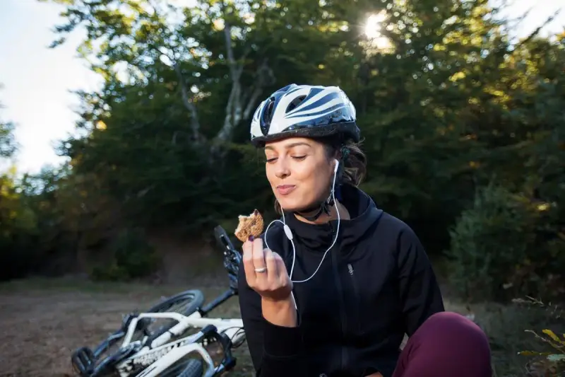 Kobieta w kasku i słuchawkach je ciastko, obok leży rower. To przykład zdrowego odżywiania i aktywnego stylu życia, czyli zbilansowana dieta.