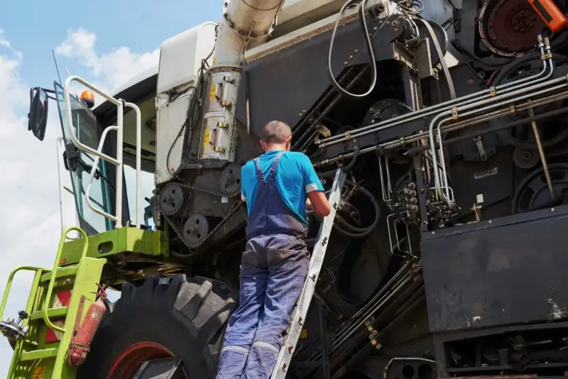 Mechanik na drabinie naprawia kombajn. Co to jest tuz? To potężna maszyna rolnicza, która zbiera plony.