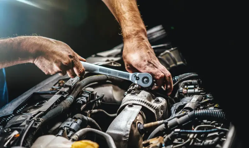 Mechanik dokręca śrubę kluczem w silniku samochodu. Widać jego dłonie i fragmenty silnika, być może podczas rutynowej kontroli lub naprawy.