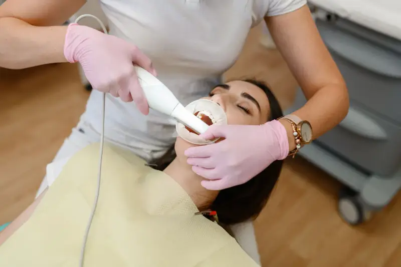 Dentystka w różowych rękawiczkach wykonuje zabieg piaskowania zębów. Czy piaskowanie zębów boli? Pacjentka ma otwarte usta i rozwierak.