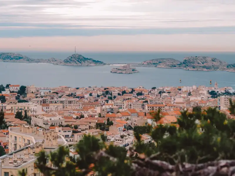Widok na miasto z dachami i morzem w tle, z widocznym zamkiem na wyspie. To piękne **Marseille**.