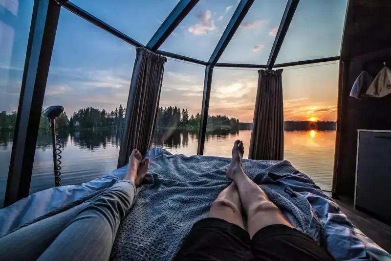 Två personer vilar i en glaskupa med utsikt över en sjö i Norrland vid solnedgången. En perfekt glampingupplevelse.