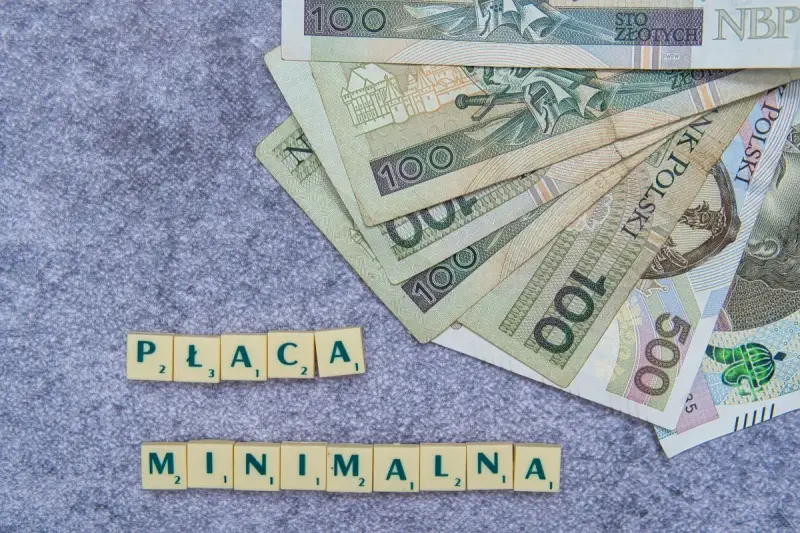 Płaca minimalna w Polsce – polskie banknoty o nominałach 100 i 500 zł ułożone na szarym tle, obok kafelki z literami tworzące napis "PŁACA MINIMALNA".