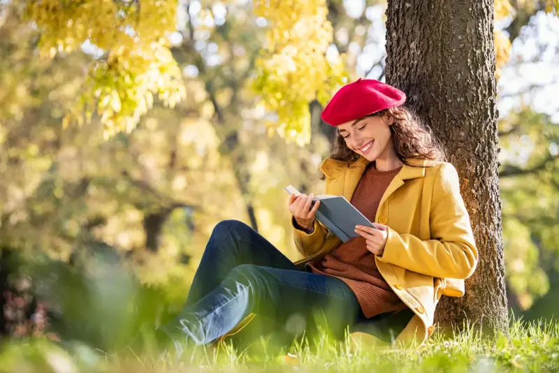Mujer con boina roja lee un libro bajo un árbol, inspirada por los temas de cuentos del otoño.