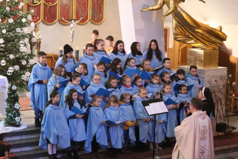 Dzieci w błękitnych strojach tworzą schola, śpiewając w kościele przy choince.