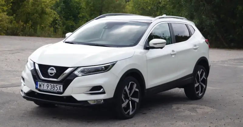 Biały Nissan Qashqai 2014 z polskimi tablicami rejestracyjnymi stoi na drodze, otoczony zielenią drzew.
