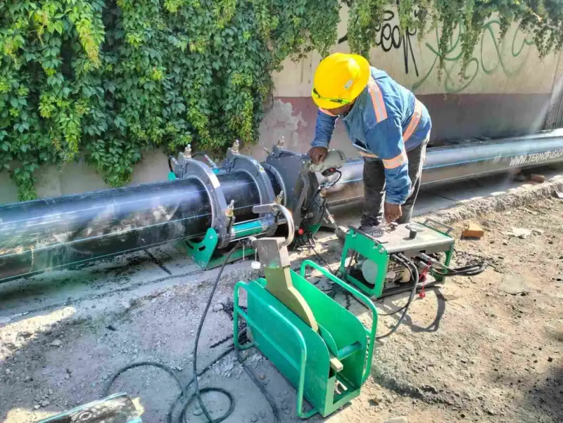 Un ouvrier utilise une machine de soudure pour joindre un gros tuyau noir. L'équipement comprend une douille à souder et une unité de contrôle verte.