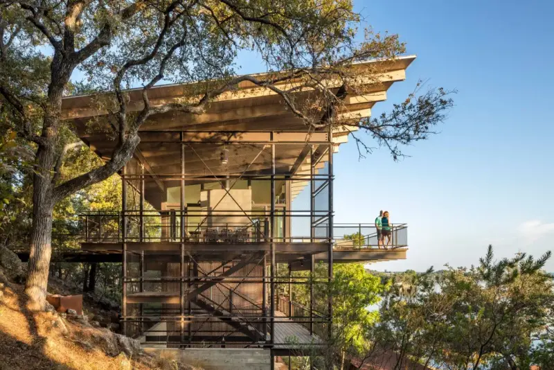 Maison moderne sur un terrain en pente raide, avec des balcons et des escaliers extérieurs en métal, nichée dans les arbres près d'un lac.