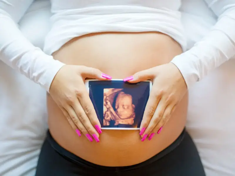 Ręce z różowymi paznokciami obejmują brzuch, na którym widnieje zdjęcie z 8 tygodnia ciąży usg 3d.
