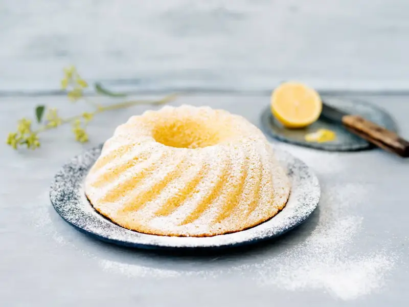 En saftig sockerkaka citron, pudrad med florsocker, serveras på ett fat. En halv citron och en kniv syns i bakgrunden.