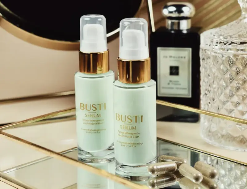 Dwa flakoniki serum ujędrniającego do biustu Busti, kapsułki i eleganckie flakony w tle.