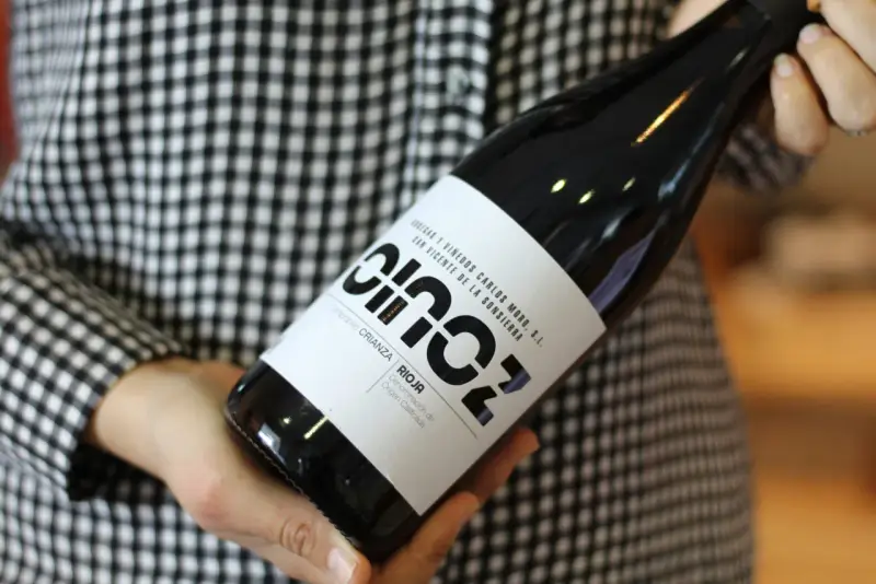 Manos sostienen una botella de vino blanco fermentado en barrica, con etiqueta negra y blanca, sobre un fondo de cuadros.