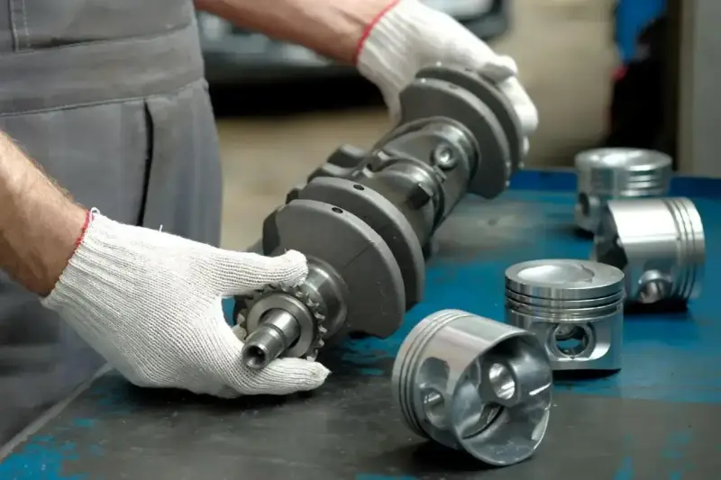 Mechanik w białych rękawiczkach trzyma wał korbowy, obok leżą tłoki. Uszczelniacz na wale jest kluczowy dla silnika.