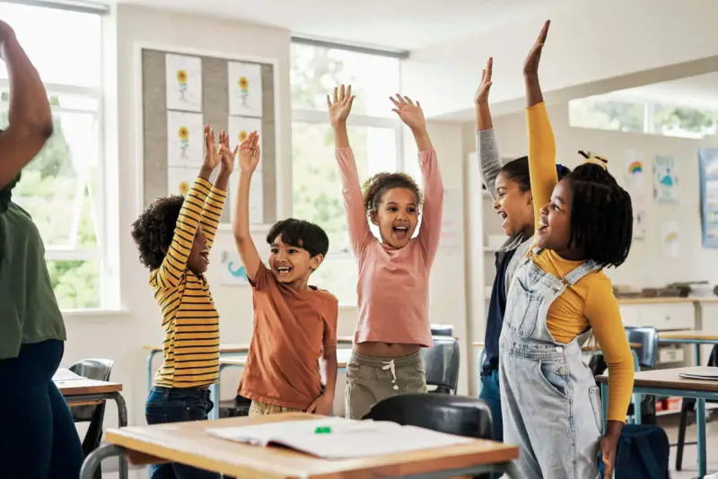 Fröhliche Kinder mit erhobenen Armen in einem Klassenzimmer. Sie nehmen begeistert an Kennenlernspielen für die Grundschule teil.