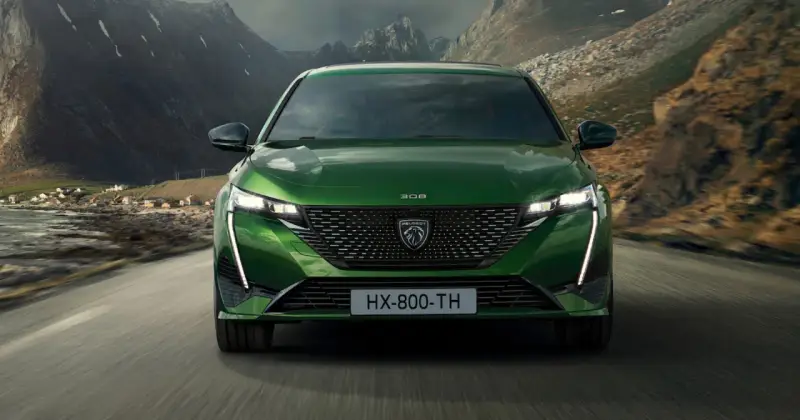 Zielony Peugeot 308 T7 jedzie drogą w malowniczym, górskim krajobrazie z widokiem na małą wioskę nad wodą.