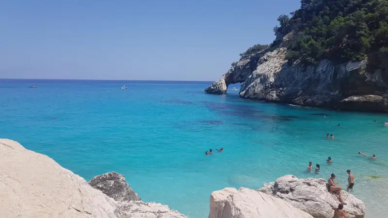 Acqua cristallina e scogliere a picco a Cala Goloritzè, raggiungibile con un suggestivo **percorso a piedi**. Persone nuotano e si rilassano.