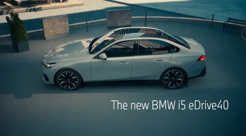 Jasnoszary sedan BMW i5 eDrive40 stoi na zewnątrz, z widokiem na basen i ławkę.
