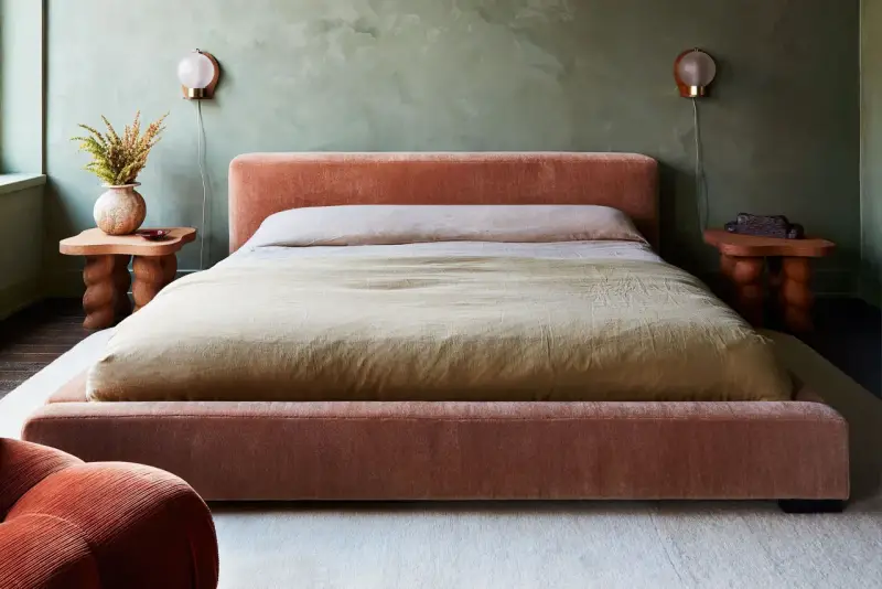 Cama king size tapizada en terciopelo rosa, con mesitas de noche de madera y lámparas de pared.