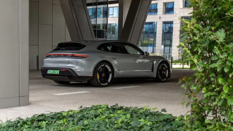 Szare Porsche Taycan Turbo S stoi pod zadaszeniem, z widokiem na nowoczesny budynek i zieleń.