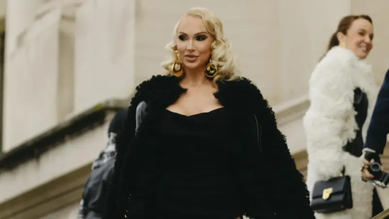 Eine Frau im Stil einer "mob wife" mit blonden Locken, großen goldenen Ohrringen und einem schwarzen Pelzmantel.
