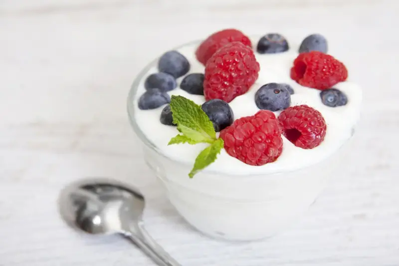 Pyszny jogurt z malinami i borówkami, idealny na śniadanie lub jako element diety jogurtowej. Obok leży łyżeczka.
