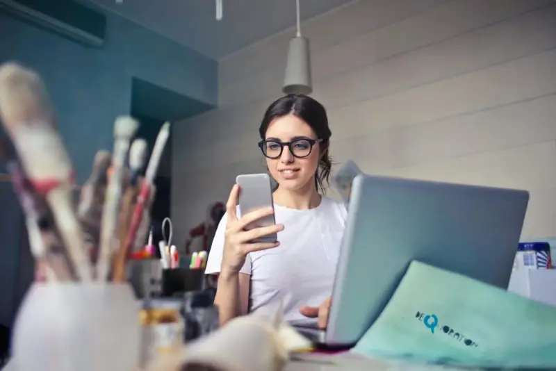 Kobieta w okularach do komputera przegląda telefon, siedząc przy laptopie.
