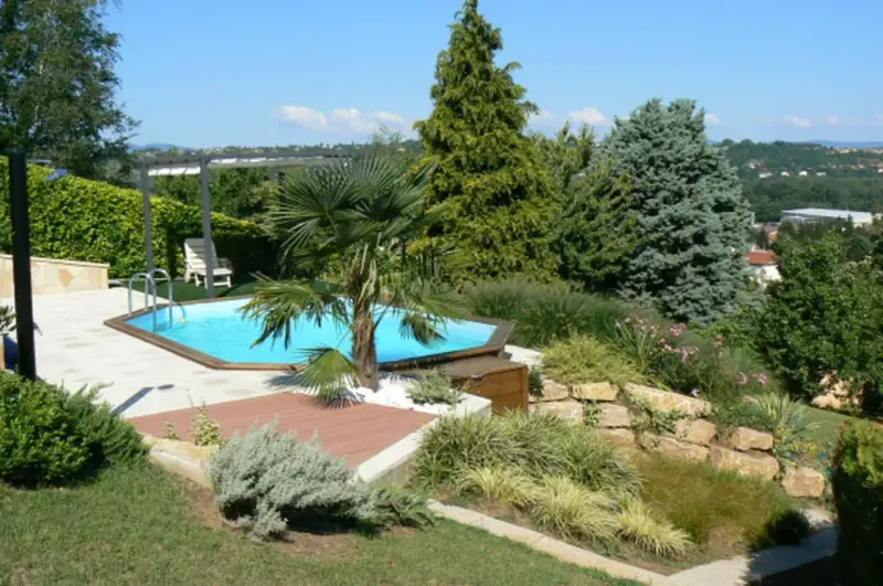 Piscine aux formes douces sur un terrain en pente, entourée de végétation luxuriante et d'une terrasse en bois.