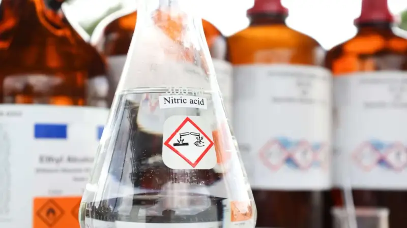 Ein Becherglas mit Salpetersäure (HNO3) und einem Gefahrensymbol. Im Hintergrund stehen weitere braune Glasflaschen mit Chemikalien.