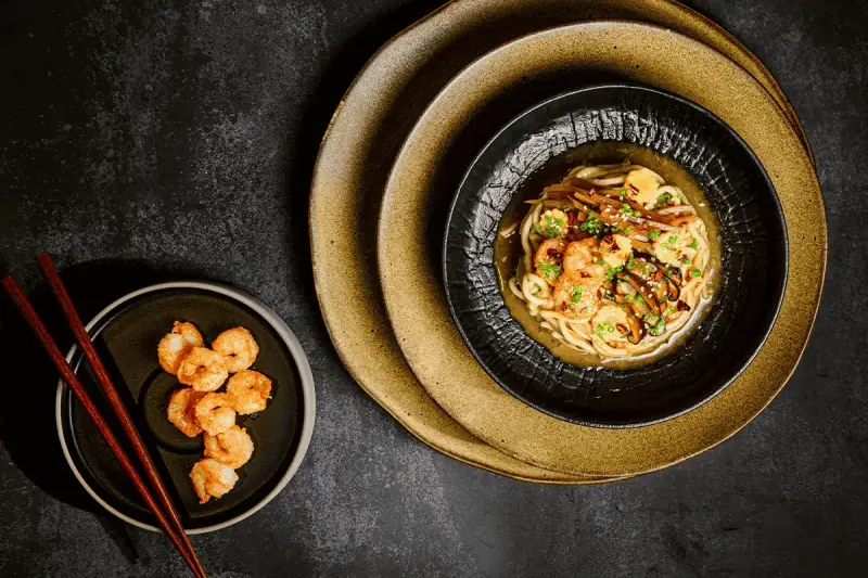 Chintan ramen z krewetkami i warzywami, podany na czarnym talerzu, obok miseczki z dodatkowymi krewetkami i pałeczkami.