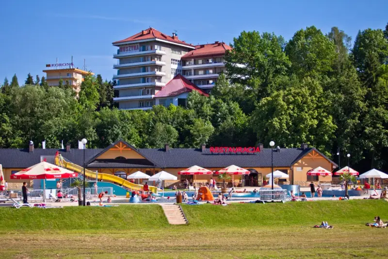 Słoneczny dzień w Muszynie-Zdroju. Ludzie relaksują się przy basenie zjeżdżalnią, a w tle widać hotel i restaurację.