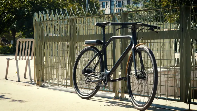 Un vélo angell noir, au design épuré, est garé près d'une grille métallique. Une chaise blanche est à gauche.