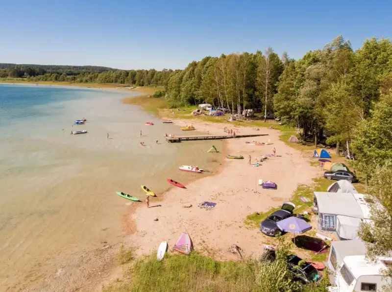 Letnie popołudnie nad jeziorem Świętajno. Ludzie kąpią się, pływają kajakami i odpoczywają na plaży wśród namiotów i kamperów.