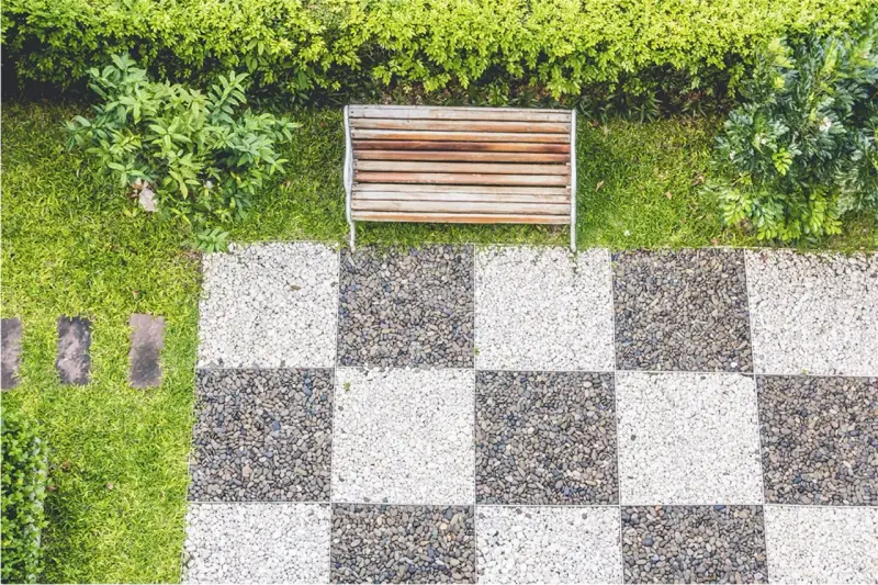 Ideas de suelo para terraza: un banco de madera sobre césped y un patrón de tablero de ajedrez de guijarros blancos y grises.