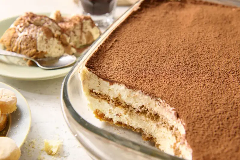 Pyszne tiramisu bez jajek, obficie posypane kakao, w szklanej formie. Obok porcja na talerzyku.