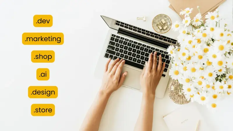 Kobieta pisze na laptopie, obok bukiet margaretek. Widoczne są różne rozszerzenia domen, np. .dev, .marketing, .shop, .ai, .design, .store.