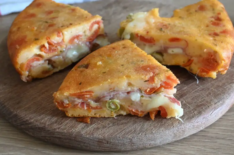 Fette di torta salata senza uova, ricca di pomodoro, formaggio filante e prosciutto, su un tagliere di legno.