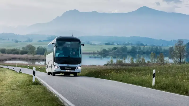 Biały **bus** marki **MAN** jedzie krętą drogą nad jeziorem, w tle góry.