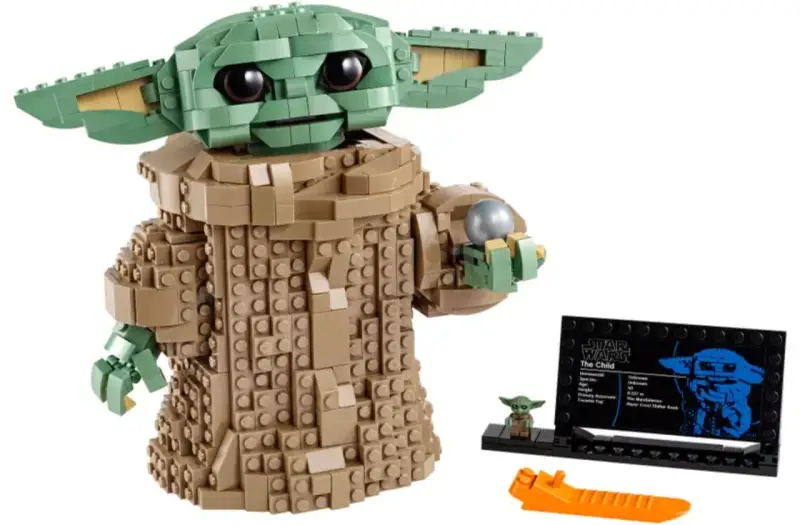 Figurka Grogu z klocków LEGO, znana z gier LEGO Star Wars, trzyma srebrną kulę. Obok stoi mała figurka i tabliczka informacyjna.