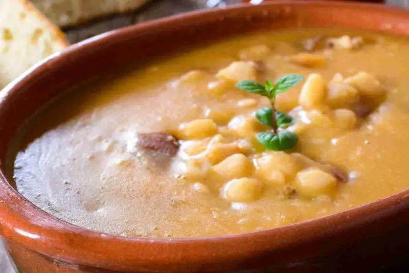 Una ciotola di terracotta piena di zuppa di fagioli cremosa, con pezzetti di carne e una fogliolina di menta. Perfetta per una cena confortante.