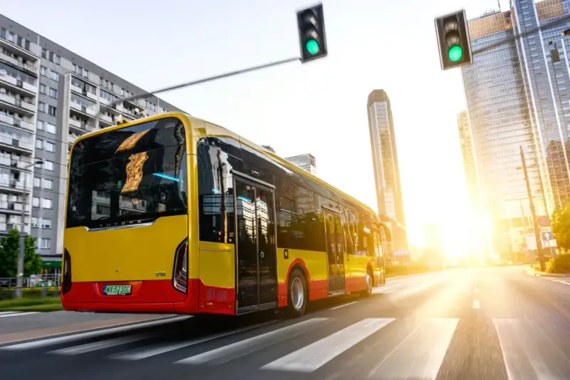 Żółto-czerwony autobus elektryczny Yutong jedzie przez miasto w blasku zachodzącego słońca.